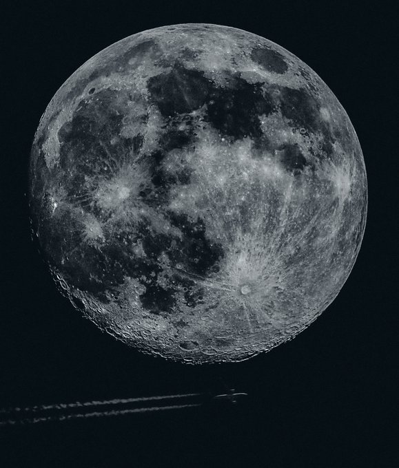 A black-and-white photograph of a detailed full moon against a dark sky, with faint airplane contrails visible at the bottom.