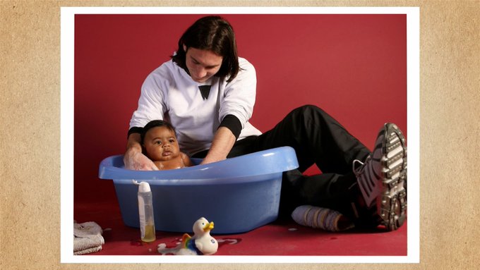 A photograph showing a person bathing a baby in a blue plastic tub against a red background, with bath accessories like a rubber duck visible nearby.