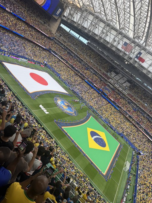 An aerial view of a stadium with large flags of Japan and Brazil displayed on the field, indicating an upcoming match between the two nations.