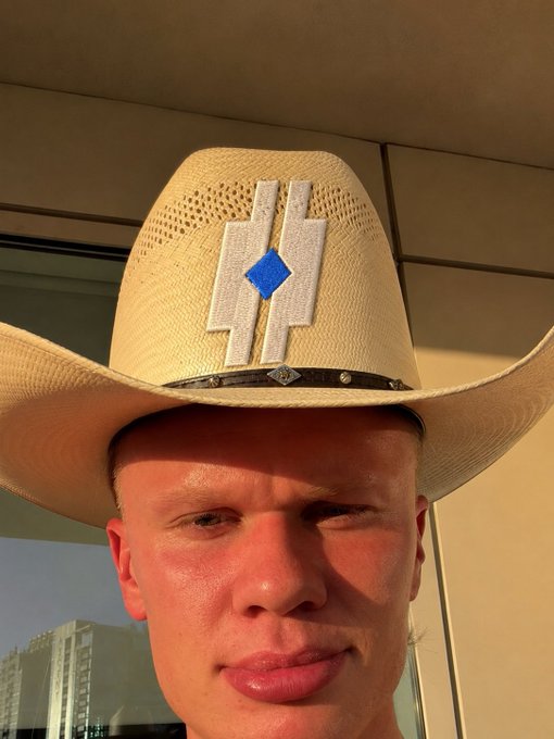 A close-up selfie of a man wearing a straw cowboy hat featuring an 'H' emblem with a blue diamond center, set outdoors with buildings visible behind him.