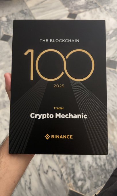 A hand holds a black and gold award plaque reading 'THE BLOCKCHAIN 100 2025, Trader, Crypto Mechanic' with the Binance logo at the bottom, against a marble background.