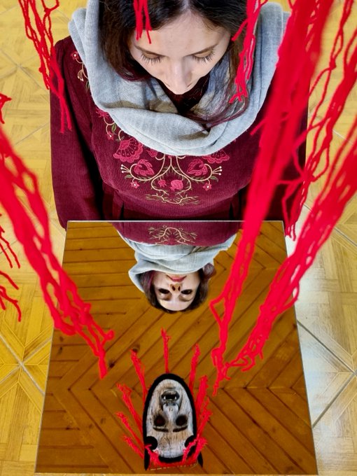 An artistic self-portrait showing a person in a maroon embroidered top looking down at a mirror that reflects their face, with red yarn/threads draped around creating a striking composition.