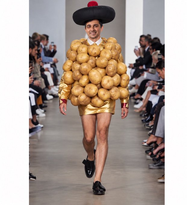 A male model walks a fashion runway wearing an avant-garde outfit consisting of a jacket covered entirely in potatoes, a black wide-brimmed hat with a red pom-pom, and a shiny gold mini skirt.