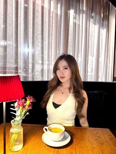 A woman with long wavy hair wearing a white halter top seated at a restaurant table with a bowl of soup, a small vase of pink flowers, and a red lamp in the background.