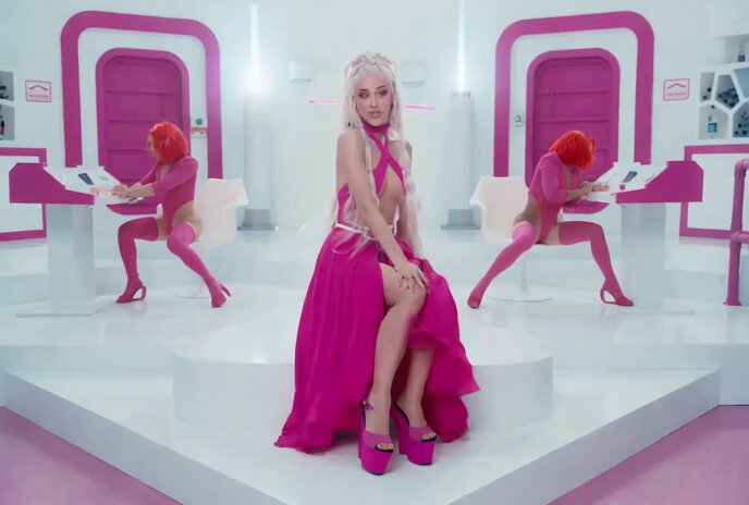 The image features a woman wearing a pink dress, sitting on a stage with her legs crossed. She has blonde hair styled in an updo. In front of her are two other women dressed in pink outfits, one standing and one sitting down. The background includes a pink room with white walls and a pink door. The caption to the image reads "Proton Pussy #IFKYK".