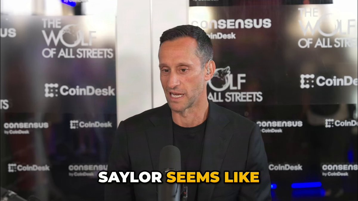 The image features a man wearing a black suit standing at a microphone, talking about his thoughts on cryptocurrency. The background of the image shows a sign that says "The Wolf of All Streets" with a logo for CoinDesk. The caption below the image reads "Saylor seems like", indicating that Saylor is being compared to the character from the movie "The Wolf of All Streets".