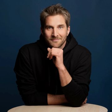 A man with short brown hair and a beard, wearing a black hoodie, sits at a round wooden table with his hand resting near his chin against a dark blue background, gazing thoughtfully at the camera. The image accompanies an article about Ledger CTO Charles Guillemet's warning that AI has shifted the advantage to attackers in crypto security, with his pointed critique of "attention farming" bug disclosures that exploit others' risk for engagement.
