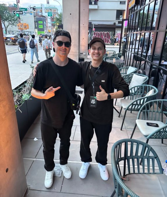 Two men posing outdoors on a city sidewalk; the man on the right wears an 'idle magic' lanyard and gives a thumbs-up, while the man on the left wears sunglasses and a black cap.