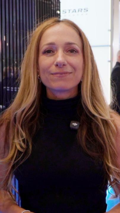 A woman with long brown hair wearing a black sleeveless turtleneck top smiles at the camera, with a DJI microphone clipped to her shirt.