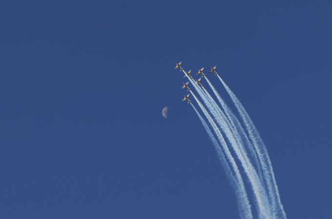 Six jets fly in tight formation, leaving long white contrails against a deep blue sky near a crescent moon.
