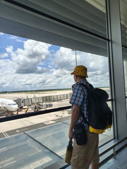 A person wearing a yellow bucket hat, plaid shirt, and backpack stands at an airport terminal window, looking out at an airplane parked at the gate under a cloudy sky.