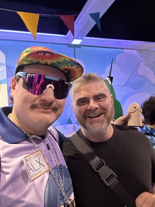 A selfie of two men at what appears to be a conference, with the man on the left wearing a tie-dye cap and purple Oakley sunglasses with a name tag reading 'ALX', posing alongside a bearded man.