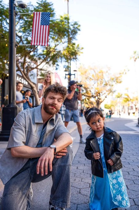 The image shows a man sitting down next to a young girl who is standing, both of them posing for a picture. The background includes an American flag flying high above their heads, indicating that they are likely in a public space with some patriotic significance or celebration. The caption on the image reads "I don't know how long we will keep pretending that all is well in this space. We need some stimmy soon to keep up the steam Am ending the day with @wallchain in my books".