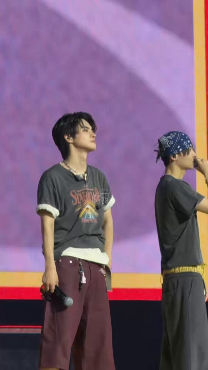 The image features two young men standing next to each other, both wearing bandanas on their heads. One of them has a microphone in his hand, suggesting that they might be performing or participating in an event. The background includes a purple wall with a logo, and there is also a banner visible in the scene.