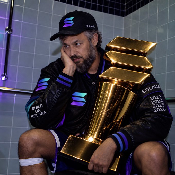 A bearded man wearing a black 'SOLANA' cap and a jacket emblazoned with 'BUILD ON SOLANA' sits on a toilet in a tiled bathroom, holding a large golden trophy engraved with the years 2023, 2024, 2025, and 2026.