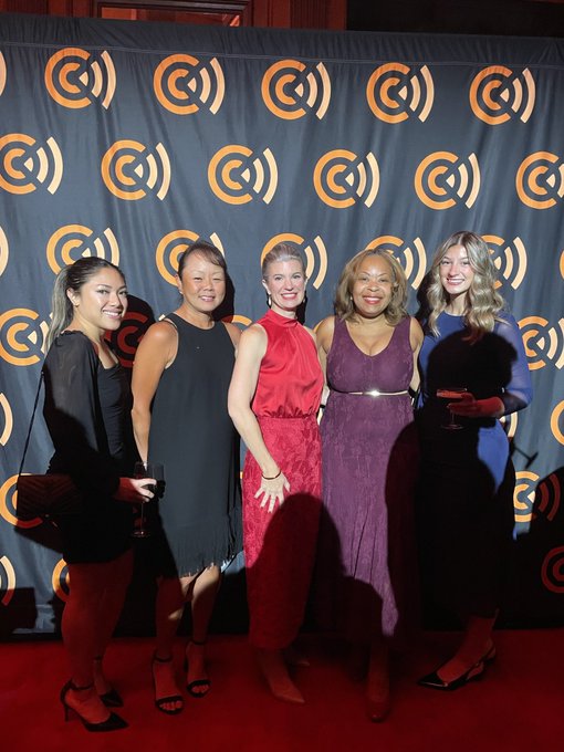 Five women pose on a red carpet in front of a black backdrop featuring a repeating orange 'C' logo pattern.