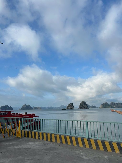 An outdoor view from a pier or dock with yellow and black striped barriers, looking out over a calm body of water with karst limestone formations in the background and a partly cloudy sky above.