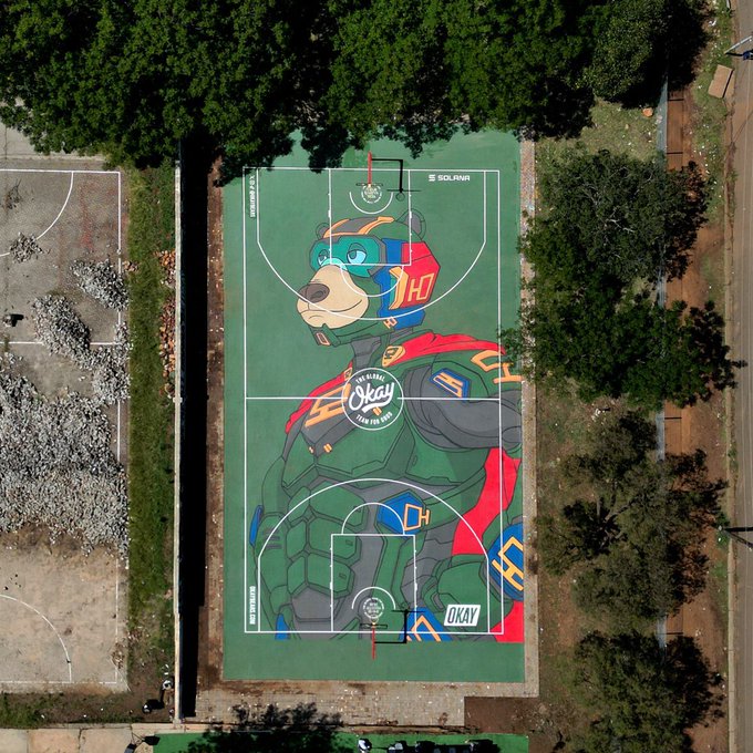 An aerial view of a basketball court featuring a large colorful mural of a cartoon dog character in futuristic gear, with 'GLOBAL Okay 5 de JUN' written in the center.