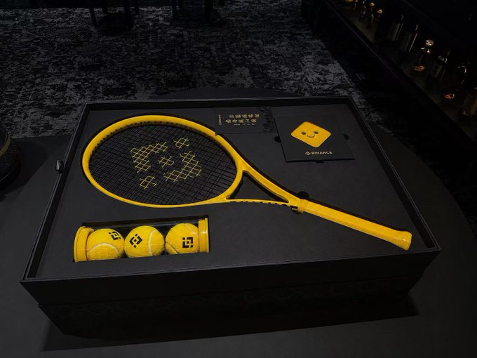 The image features a tennis racket, three yellow tennis balls, and a box containing two more tennis rackets inside of a black box. The tennis ball packaging includes a smiley face design. The scene takes place against a black background with white text that reads "@binancezh 的端午周边展览馆了，盒子巨大，龙鳞花纹帅爆改币圈周边展览馆".