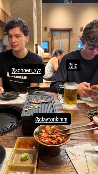 Two young men, tagged @schoen_xyz and @jiz, sit at a Japanese restaurant table enjoying a meal of grilled meat and kimchi, with @claytonkimm filming the scene. A large mug of beer sits between them as they share the dining experience at what appears to be a yakiniku establishment.