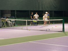 Four people stand on a purple indoor tennis court near the net, with chairs and equipment visible along the sidelines.