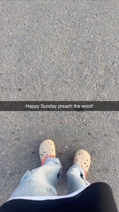 A first-person view of feet in white Crocs and light blue jeans standing on asphalt, with a text overlay reading "Happy Sunday preach the word!".