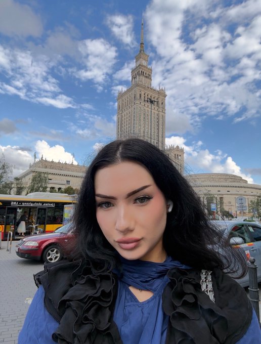 A woman with long black hair poses in front of the Palace of Culture and Science in Warsaw, with city buses and cars behind her.