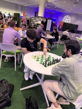 The image shows a group of people gathered around tables, playing chess. There are at least six individuals visible in the scene, with some sitting on chairs while others stand or sit on the floor. The main focus is on two men engaged in a game of chess, with one man making his next move and the other attentively observing the board.

The setting appears to be an indoor space, possibly a convention center or a similar venue. There are multiple tables spread across the room, each hosting a player participating in the game. A backpack can also be seen placed on the floor near one of the tables. The overall atmosphere suggests that this is a social event where people have come together to enjoy playing chess and engaging with others who share their interest in the game.