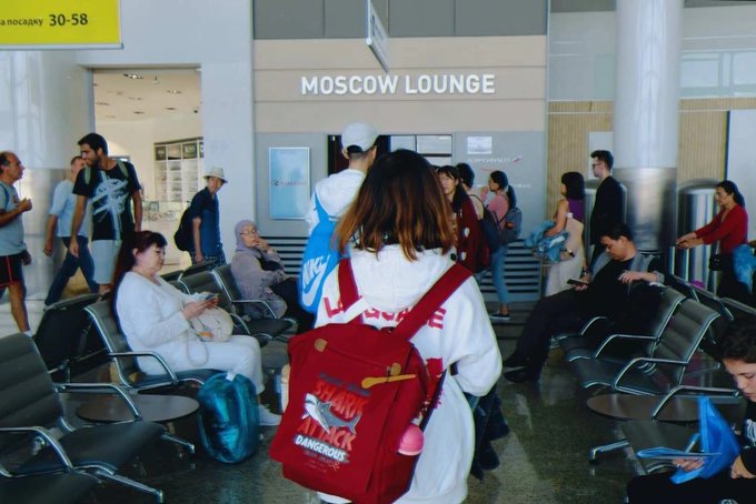 Travelers sit and walk in an airport terminal near a 'MOSCOW LOUNGE' entrance, with a foreground figure carrying a red backpack.