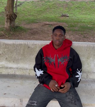 The image features a young man sitting on a concrete bench, wearing a red hoodie with black designs. The man has his hands together and appears to be posing for the photo. The background of the image shows a grassy area next to some trees, creating an outdoor setting. There is also text in the image that reads "Fresh cut to predict better with @BitgetWalletAF Rate my haircut I went from this. To this". This suggests that the man might have recently gotten his haircut and wants to share his experience or recommend a hairstylist for others.
