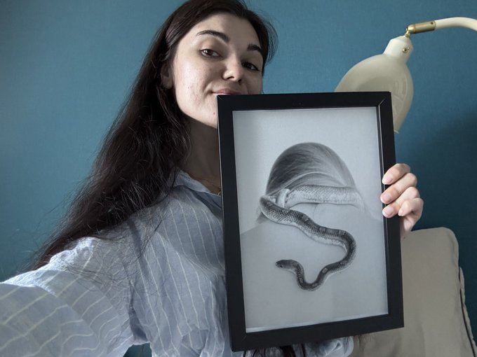 A smiling woman with long dark hair holds up a framed grayscale drawing of a snake's head in front of her face, with a white lamp visible in the background.