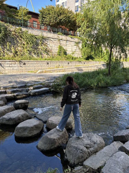 A person wearing a dark hoodie with "WORLD WIDE" text and a bear graphic stands on stepping stones crossing a shallow stream, with stone walls, ivy, and willow trees lining the urban waterway. The image captures a moment of casual exploration en route to a pop-up event, embodying the "OTW to the Pop-Up" vibe.