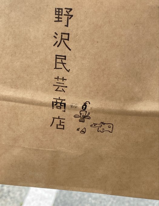 A brown paper bag with Chinese characters 野沢民芸商店 (Nozawa Mingei Shoten) printed vertically, along with a small stamped illustration of vegetables and a figure.