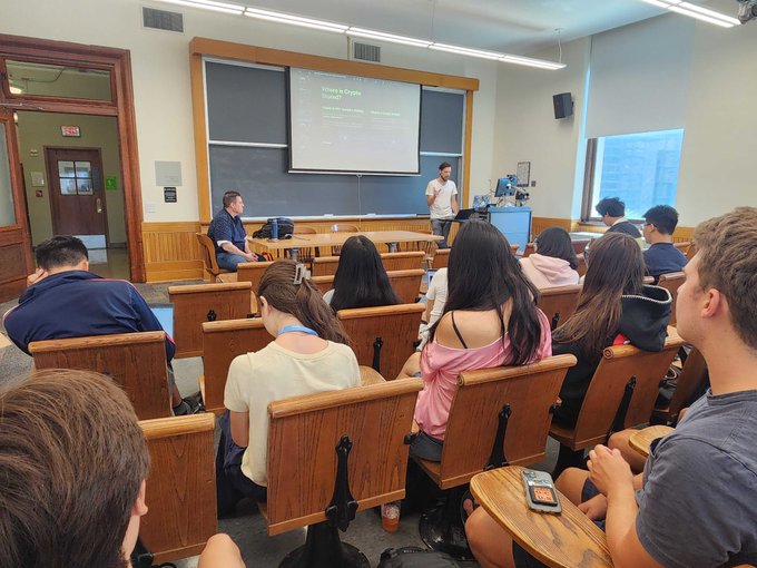Students sitting in wooden desks in a lecture hall attending a class presentation displayed on a projection screen.