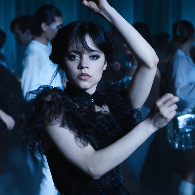Jenna Ortega stars as Wednesday Addams in a dark, moody scene, dancing with her arms raised in a black ruffled dress surrounded by blurred figures.