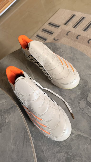 A pair of sleek white soccer cleats with vibrant orange accents rests on a marble surface, ready to hit the field as a well-earned reward for the buyer.