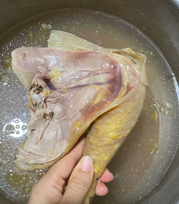 A hand holds a raw, partially butchered chicken leg quarter over a metal basin containing murky water and oil droplets.