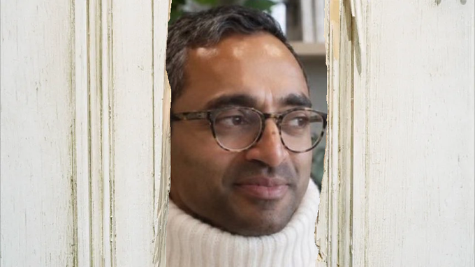 A man with glasses and a dark beard, wearing a cream-colored turtleneck sweater, peeks through a narrow gap in weathered white wooden slats. The playful, peering pose aligns with the post's theme of "crashing" an event, capturing the mischievous spirit of sneaking into a Federal Reserve gathering to discuss crypto.