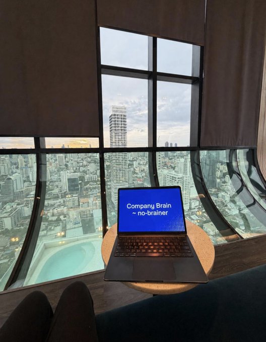 A laptop displaying the text "Company Brain – no-brainer" sits on a stool by a curved window overlooking a sprawling cityscape, visually reinforcing the post's pitch that a $100/month AI "company brain" service is a steal compared to the premium office setting suggested by the high-rise view.