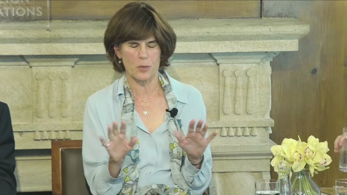The image shows a woman sitting at a table with her hands up, possibly gesturing or expressing her thoughts during a discussion. The setting appears to be an event where people are gathered around tables, likely engaged in conversation about various topics. There is also a vase of flowers on one side of the room and a chair nearby.