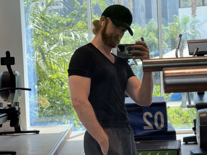 A man with a reddish beard, wearing a black cap, white earbuds, and a black t-shirt, taking a mirror selfie with his phone in what appears to be a gym.