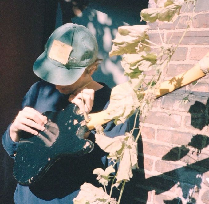 A vintage-style photograph of a person wearing a faded green bucket hat and dark sweatshirt, playing a black electric guitar while standing among plants near a brick wall, with sunlight filtering through leaves.