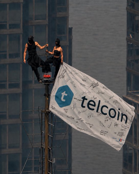 Two people in black balaclavas stand on a tall tower mast displaying a 'telcoin' flag covered in signatures, with high-rise buildings in the background.