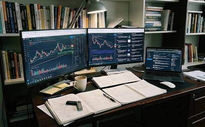 A trader's desk setup shows two large monitors displaying candlestick charts and analytics, a laptop showing data, an open notebook with handwritten calculations, a coffee mug, and bookshelves filled with books in the background.