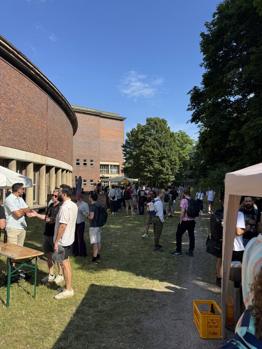 The image shows a large group of people gathered outdoors around tables with umbrellas, possibly at an event or gathering. The crowd consists of men and women standing close together, engaged in conversation and enjoying each other's company. There are also several backpacks visible among the attendees. In the background, there is a building that appears to be made of brick, which could be a venue for the event taking place.