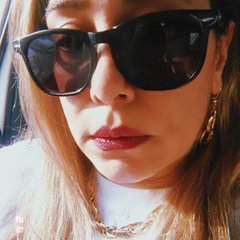A close-up selfie of a woman wearing large black sunglasses, with the reflection of the photographer's hands visible in the lenses, and layered gold necklaces.