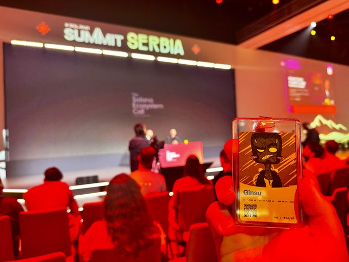 A hand holds up a phone displaying a "Ginsu" character badge at the "SUMMIT SERBIA" event, with a stage and audience visible in the background.