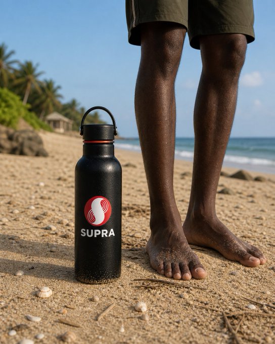 A black water bottle branded with the 'SUPRA' logo sits on a sandy beach in front of a person's legs, with the ocean and palm trees in the background.