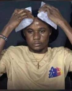 A young man in a yellow shirt holds two ice packs or cloths to his forehead with both hands, wearing a cross necklace, with a distressed or tired expression.