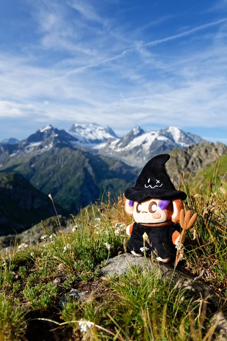 A small plush toy dressed as a witch with a black hat stands on a grassy mountain overlook with snow-capped peaks in the background.